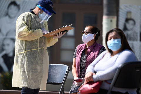 Testing for Coronavirus. A healthcare worker in protective gear, stands and holds a clipboard as two Latinx women sit with masks on  