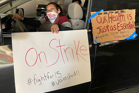 Fast food protest. Latinx woman in car wearing a face mask in black car holding a white sign that reads: On Strike #fightfor15 #unionforall