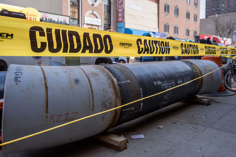 Yellow caution tape surrounds rows of pipeline left on the side of an urban street. 