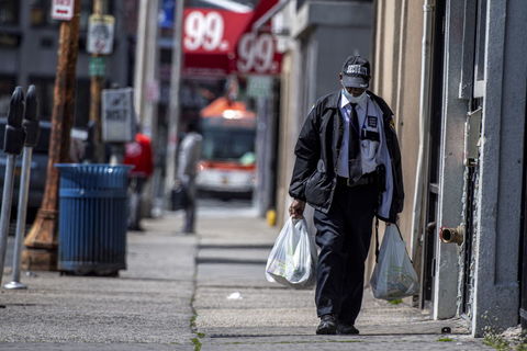 A Black man wearing a security guard uniform and a mask due to coronavirus concerns walks down a desolate New York street 