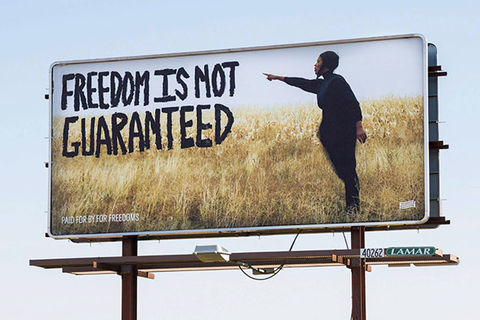 Billboard. Photo of a young Black man standing in a field of tall grass pointing to words in big black letters that read: "Freedom Is Not Guaranteed."