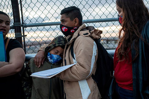 Immigration. Latinix father wearing tan coat and black face mask stands in front of gate with young boy leaning on him, also wearing a mask, and Latinx woman wearing red shirt and black jacket, with a face mask.