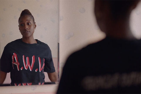 Insecure. Black woman looking in mirror at herself in bathroom, wearing black N.W.A. tee shirt.