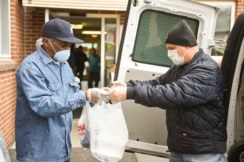 Latinx Community. Elderly man wearing blue shirt and baseball cap accepts food from the back of a white truck, where a man in black stands handing out food.