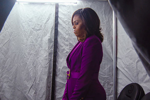 Michelle Obama. Black woman with dark shoulder length hair standing in a concentrated lit space wearing a purple suit.