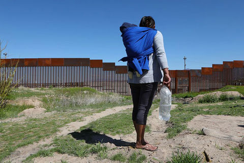 Migrant. Rear view of woman with dark hair wearing gray top, dark leggings and carrying a child in a blue blanket and a large bottle of water, facing the U.S./Mexico border wall.