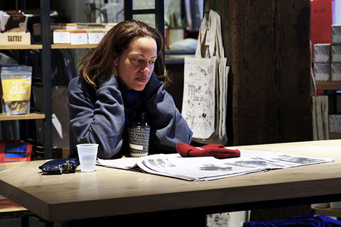 Drew Dixon. Black woman with shoulder length brown hair wearing blue sweatshirt sitting alone at table in coffee shop.
