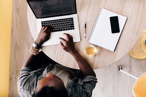 Positive Peers. Aerial shot of Black man working at a laptop with smartphone and white notepad on table, next to golden colored drink.