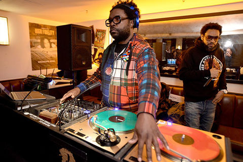 Questlove. Black man with short dark hair DJ'ing, wearing headphones, an orange and blue plaid shirt and dark framed glasses.