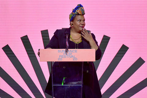 Radha-Blank. Black woman wearing colorful head wrap and dark purple dress, standing in front of podium that reads Sundance Film Festival 2020 and a pink background.