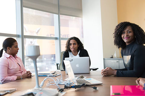 Small Business. Three women of color sit at a conference table with large windows and computers.