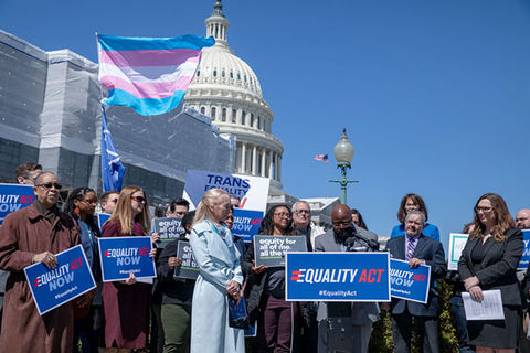 Trans health. A cluster of people in winter coats standing around a podium holding blue signs with the trans flag waving in the background in front of the U.S. Capitol.