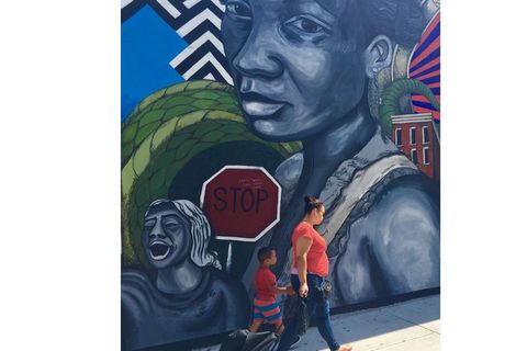 A Latinx mother and child with bright red shirts walk past wall-high mural with Black Latinx woman and child. 