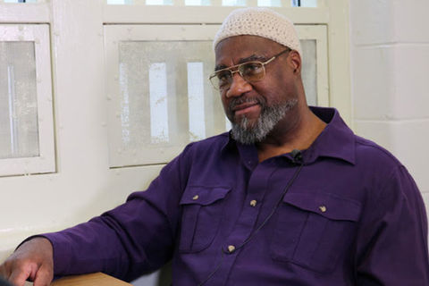 Jalil Muntaqim. Older Black man wearing white kufi and purple shirt and glasses.