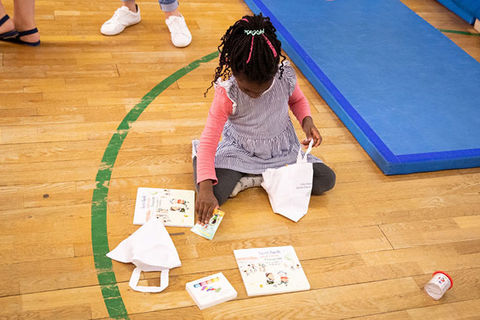 Literacy. Small Black girl with braids wearing a pink top and blue strip dress kneels on gym floor around papers.