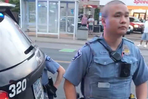 Asian police officerwith a shaven head wearing a blue uniform standing in front of a black police cruiser.