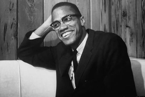 Malcolm X. Archival black and white photo of Black man wearing dark suit, white shirt and black tie.