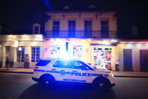 NOPD. White police cruiser with blue letters reading Police New Orleans, driving down a street with lights on.