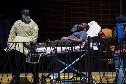 NYC-COVID-19. Male healthcare worker wearing blue baseball cap, blue face mask and yellow PPE coat wheels in a Black male patient on a stretcher who is wearing a blue Tee shirt and blue pants.