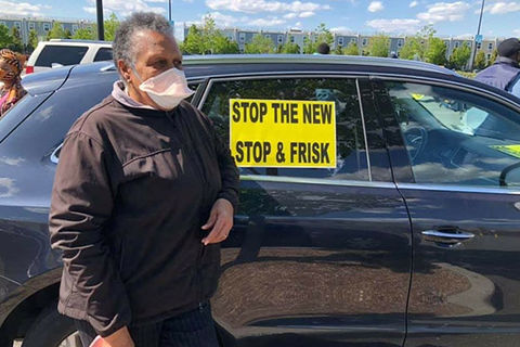 NYPD Protest. Older Black woman with short gray hair wearing white face mask and black jacket standing in front of black car with sign in window that reads: Stop the new stop & frisk.