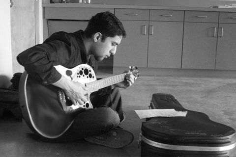 Raef. Black and white photo of man with short dark hair sitting on floor playing guitar in front of guitar case, wearing dark colored shirt and pants.