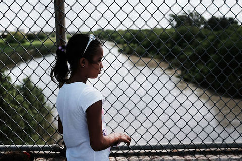 Young girl with dark hair stands in front of a wire fence that overlooks a stream of water. 