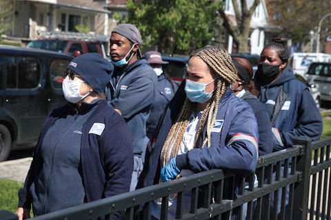A group of postal workers in uniform gather on the sidewalk to honor a fellow colleague that died of COVID-19.