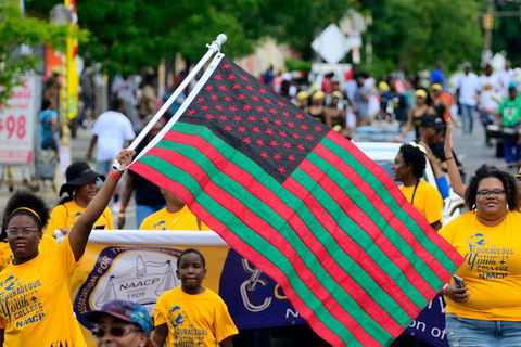 a group of young, Black people wearing yellow shirts march in a parade as they wave a red, black and green flag.