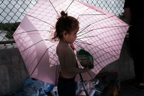 A small child stands shirtless in front a wire fence as she holds an open, pink umbrella. 
