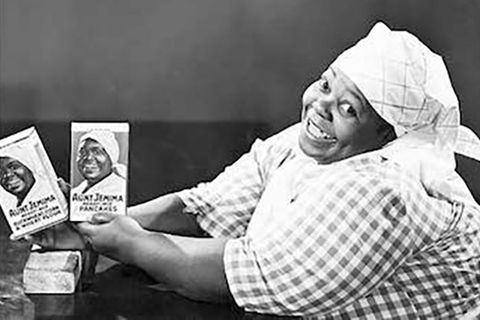 Nancy Green. Black and white image of an older Black woman wearing a white head scarf and gingham dress, holding two photos of Black women that read "Aunt Jemima."