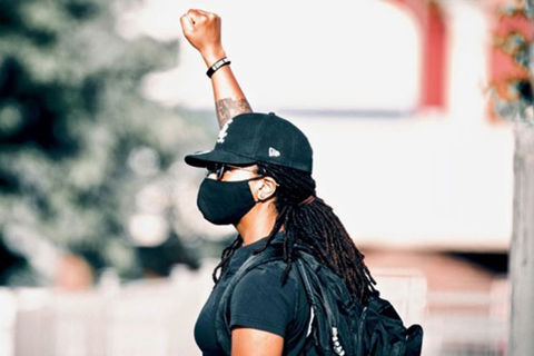 Autostraddle. Black woman wearing black cap, black face mask, black Tee and black back pack with one fist raised in the air.
