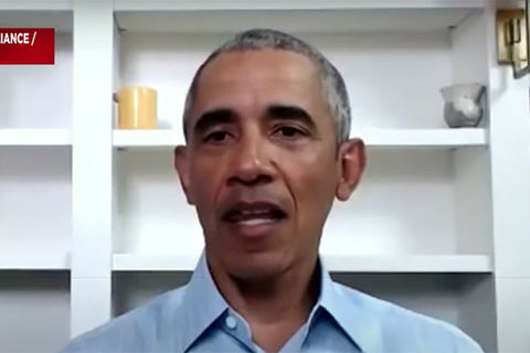 Barack Obama. Black man with short gray hair wearing a blue buttoned shirt sitting in front of a white bookshelf.