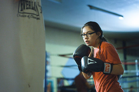 Donna Kipp. Young Blackfeet woman with dark hair, wearing glasses, orange Tee,  black boxing gloves standing in front of a body bag.