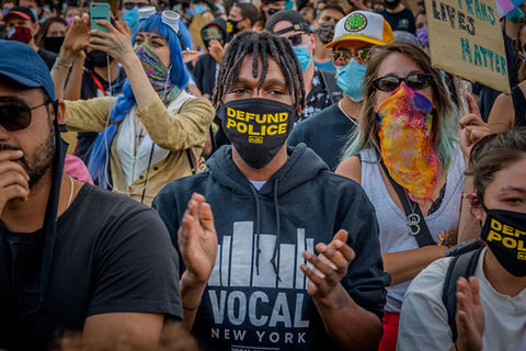 Defund Police. Black male protestor in a crowd wearing a black face mask that reads "defund police" in yellow letters, wearing a black hoodie.