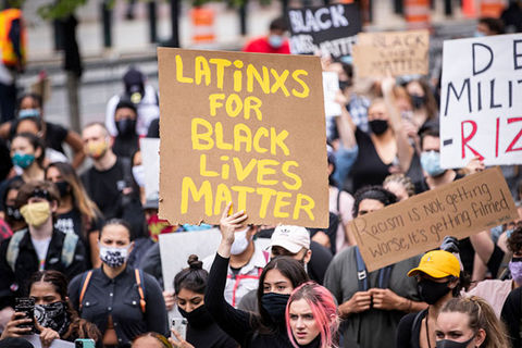 Latinx for BLM. Crowd of protestors where one woman holds up sign with yellow letters reading: "LATiNXS FOR BLACK LiVES MATTER"