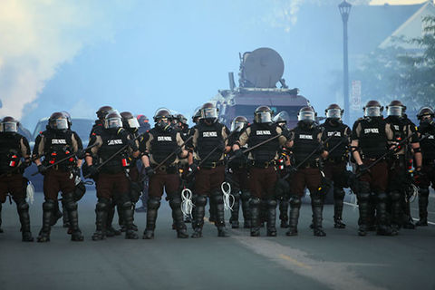 Minneapolis PD. A row of police in black uniforms wearing riot gear, standing in front of an armored vehicle.