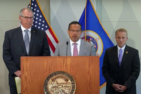 MPD Charges. Black man wearing glasses and gray suit stands in front of podium next to two White men wearing black suits.