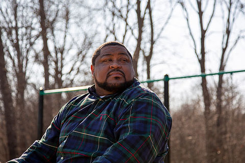 Robert Williams. Black man with short dark hair and facial hair wearing a blue plaid shirt in a park.