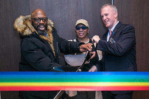 Stonewall House. Ribbon in rainbow colors being cut by Black man with bald head and dark glasses wearing dark winter coat, Black woman wearing tan cap and White man with gray hair wearing dark suit.