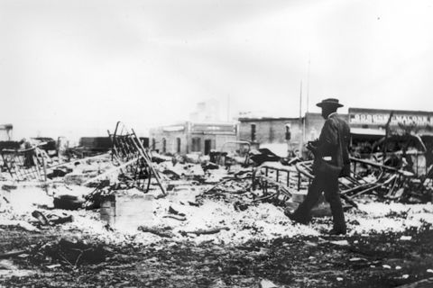An African-American man with a camera looks at the skeletons of iron beds which rise above the ashes of a burned-out block after the Tulsa Race Massacre, Tulsa, Oklahoma, June 1921