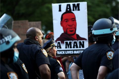 Four NYPD officers in helmets face a crowd of protesters in NYC, one with a sign of George Floyd and the words "I am a man"