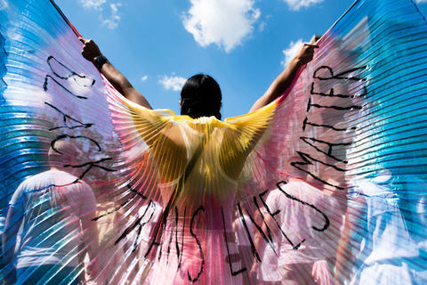 A person holding multicolored wings that read Black Trans Lives Matters outside under a blue sky.