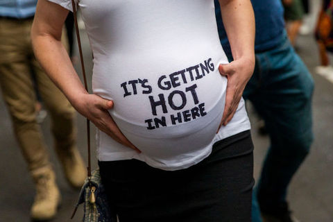 A pregnant stomach is cradled by arms and hands, in a white t-shirt that reads "It's Getting Hot in Here."