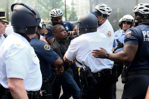 A Black man wearing a black shirt is struggling with at least seven police officers as they surround him. 