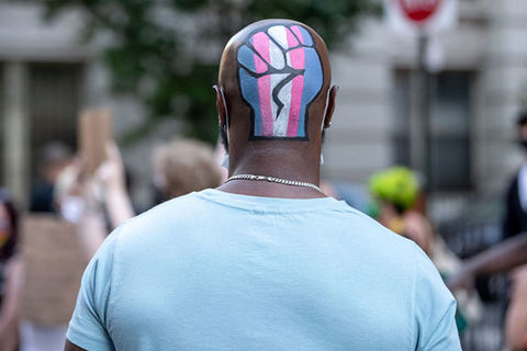Black Trans Lives. Black man with bald head wearing blue shirt has Trans Pride flag colors painted onto back of head in the shape of a fist.