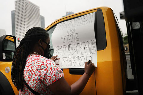Black-student. Black woman with long braids and red/white dotted shirt writes on sign, "Invest in students period."