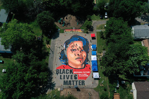 Breonna Taylor mural. Large paining on ground surrounded by trees in a neighborhood of a young Black woman with blue hair and red shirt.