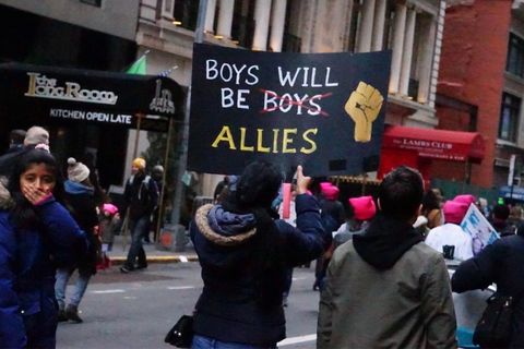 Image of a protest march where a young Brown girl is covering her mouth, and a woman has a sign reading "Boys Will be Boys Allies" with the second "Boys" crossed out
