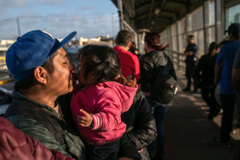 A man wearing a blue baseball cap kisses his small daughter as he holds her in his arms. 