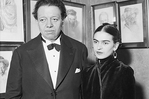 Diego Rivera and Frida Kahlo. Black and white photo of tall Latinx man in black suit with white shirt stands next to smaller Latinx woman wearing black.